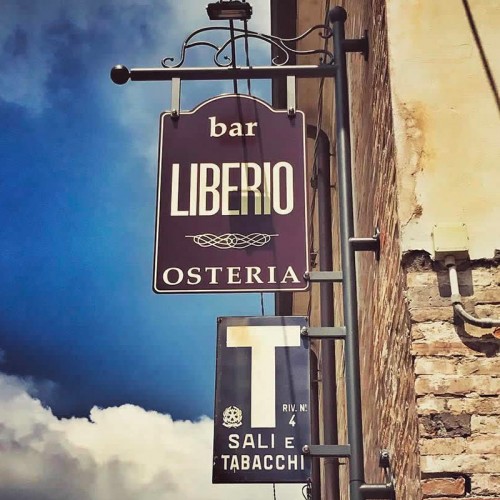 Insegna del Bar Liberio Osteria a Cesenatico, con un cielo blu e nuvole sullo sfondo, che mostra anche un cartello per la vendita di sali e tabacchi.