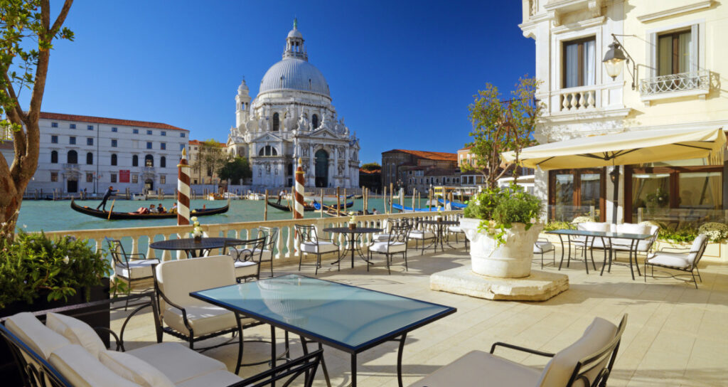 L'immagine rappresenta una terrazza sui canali veneziani! Location suggestiva che vede dei tavolini affacciati sulla laguna con una chiesa e palazzi storici sullo sfondo.