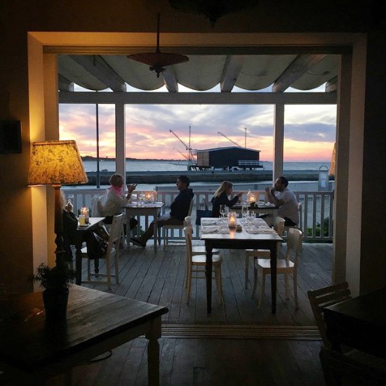Una vista suggestiva di un ristorante all'aperto durante il tramonto, con tavoli apparecchiati e candele accese. Alcune persone sono sedute ai tavoli, godendosi la vista del mare e del cielo colorato, mentre sullo sfondo si intravede un molo con delle gru. ```