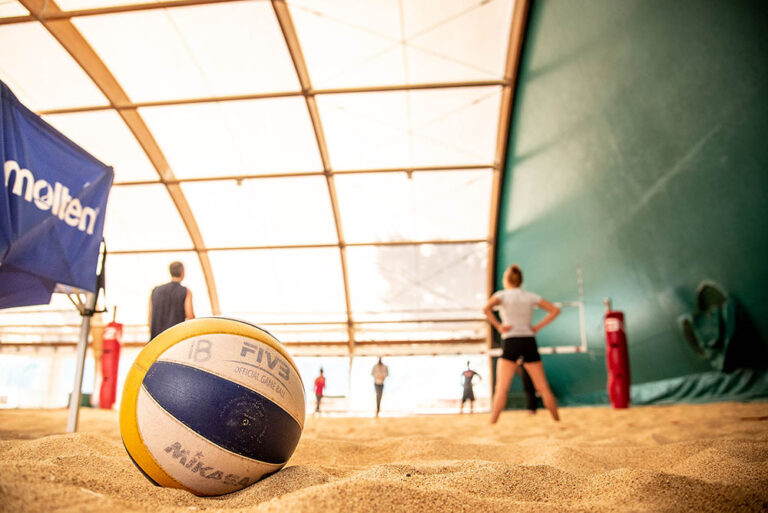 campo di beach volley con una palla in primo piano e alcuni giocatori ed in carrello per palloni più indietro sullo sfondo