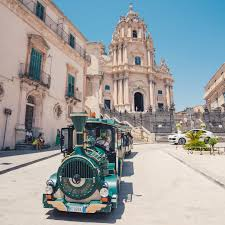 Il Trenino Barocco di Ragusa Ibla in primo piano, con il Duomo di San Giorgio sullo sfondo, immerso in un'atmosfera luminosa di una giornata soleggiata